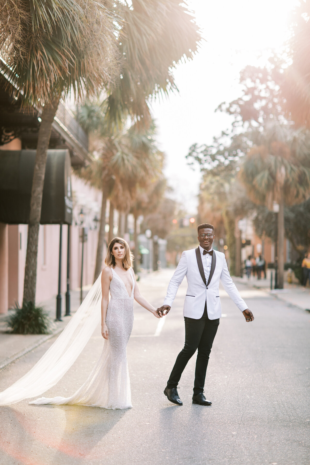 Gorgeous portrait of a young bride in a wedding gown holding the hand of a handsome groom in a white tuxedo with black lapels walking across the street in Charleston with the sunset glowing behind them