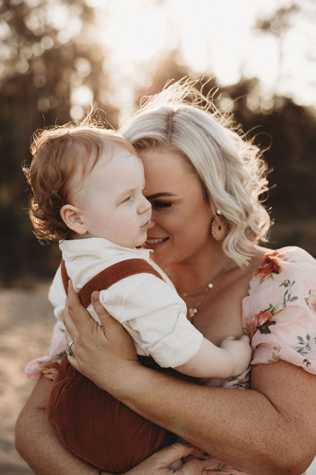 A woman with blonde hair wearing a floral dress holds a baby with red hair dressed in a white shirt and brown overalls. They are outdoors, and the woman gently nuzzles the baby's head, capturing a tender moment perfect for South Coast family photography.