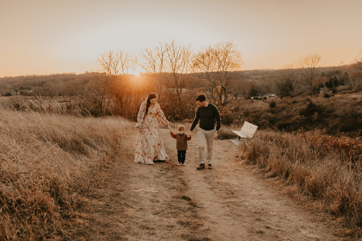 A family of three enjoys a golden hour walk along a scenic dirt path in Morris County, New Jersey. The warm sunset light highlights the tall grass and bare trees, creating a serene and picturesque backdrop. The mother wears a flowing floral dress, the father is dressed in neutral tones, and their young child is bundled in a cozy jacket. 