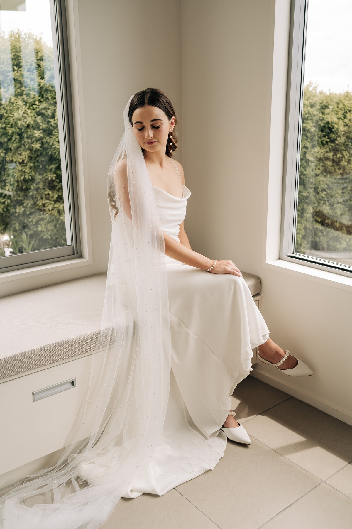 bride with white grace loves lace dress and pearl shoes and pearl veil sitting by window on wedding morning at port levy christchurch
