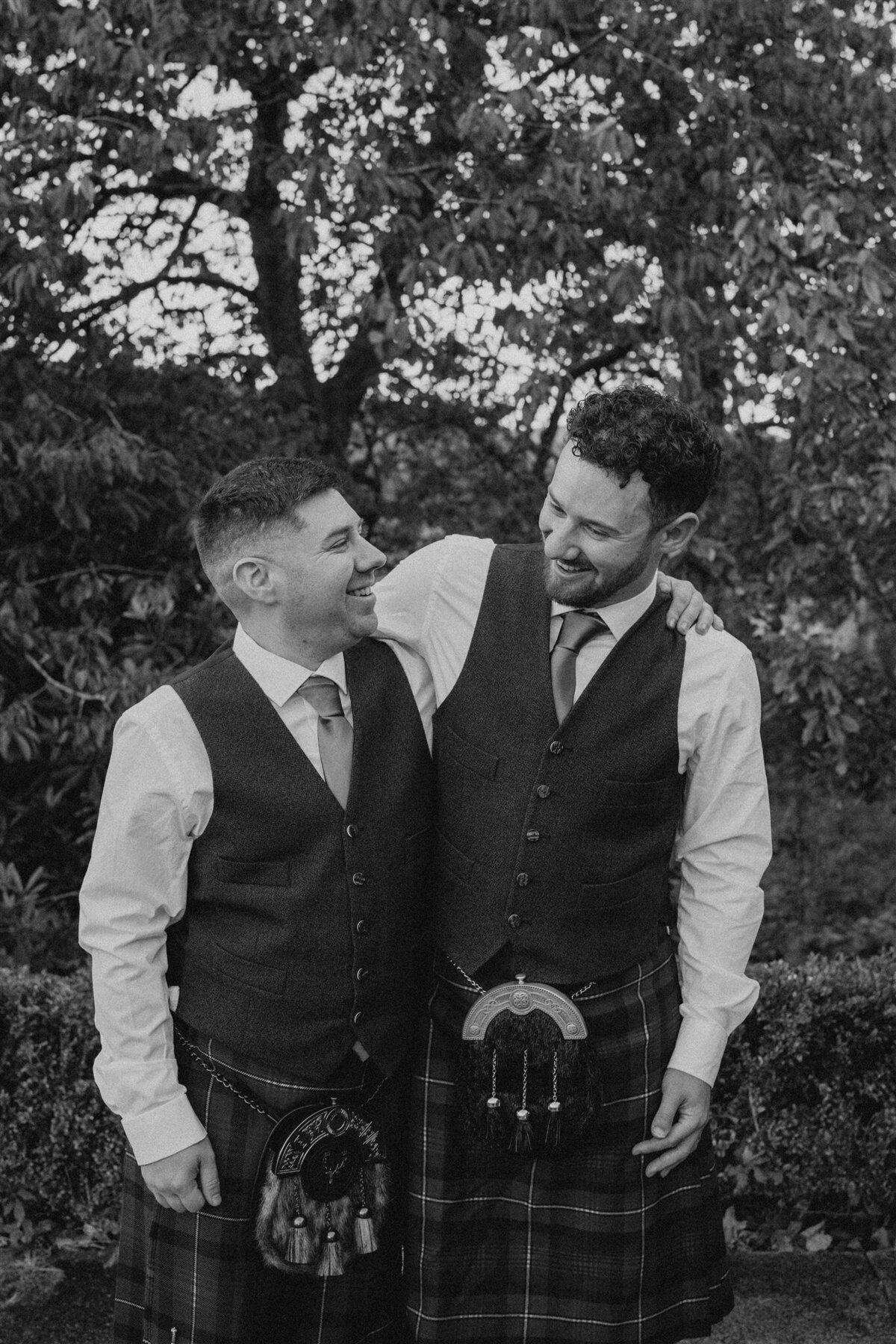 Two men stand side by side and pose for a photograph. They both wear kilts and traditional Scottish wedding attire. Trees and greenery surround them.