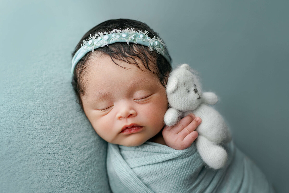 newborn baby girl in a swaddle on her back    on a sea green backdrop with matching headband and holding a white bear at a newborn photo shoot in Northern VA