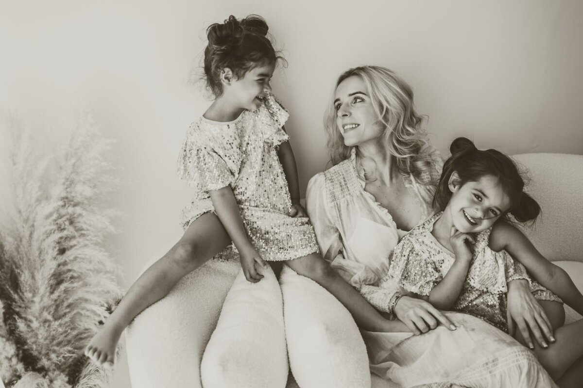 a mother with her two daughter sitting on a couch in a brownish color photo and the mom and 1 daughter smiling to each other