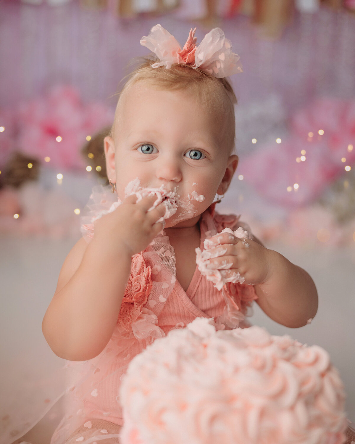 one year old baby girl with blonde hair and blue eyes wearing pink sitting in front of a frosted cake while she smashed it with her hands