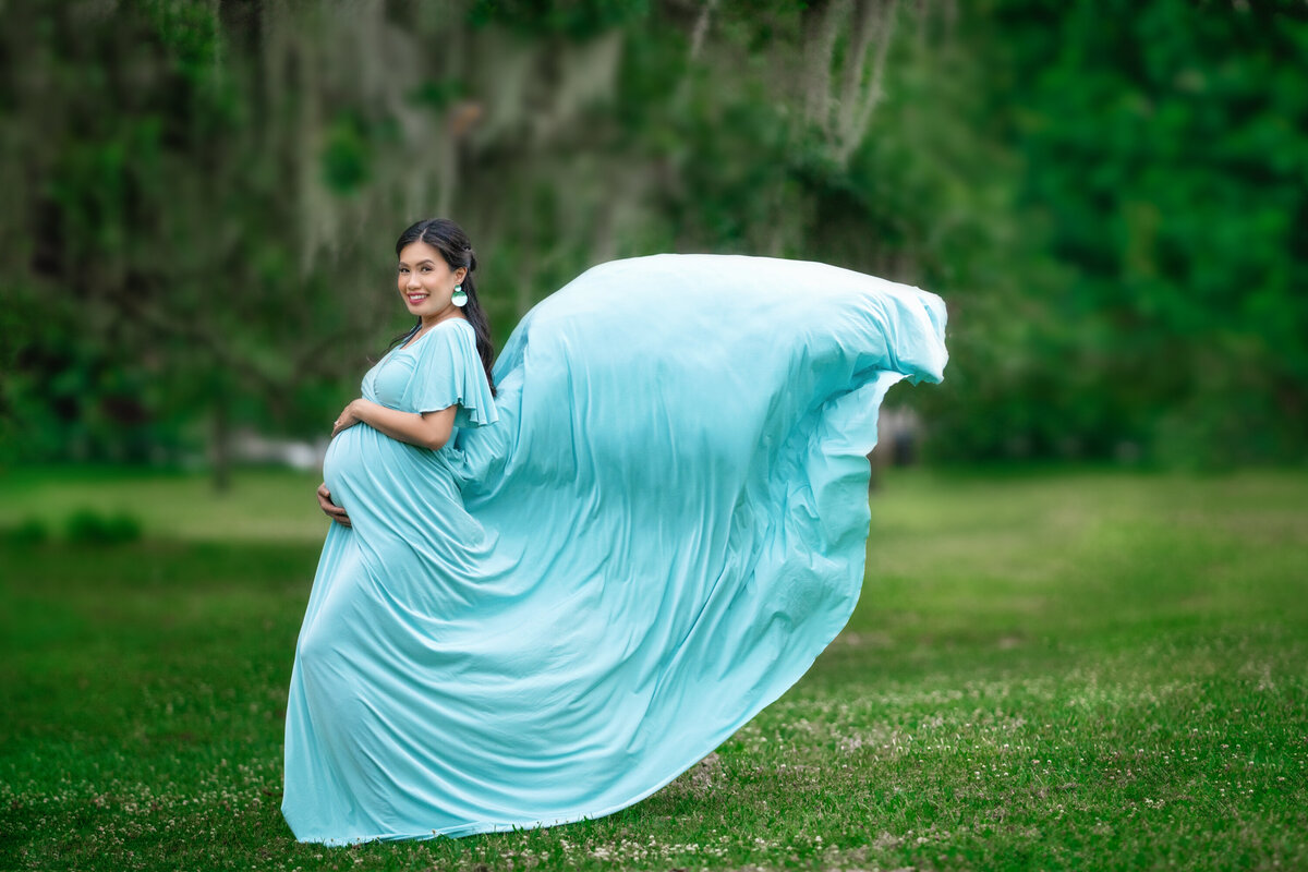 Pregnant Asian woman standing under oak trees and Spanish moss in Audubon Park.  She has black hair pulled back and a big smile.  She is holding her belly and looking at the camera.  She is wearing an aqua Silk Fairies Maternity dress which is blowing in the wind.