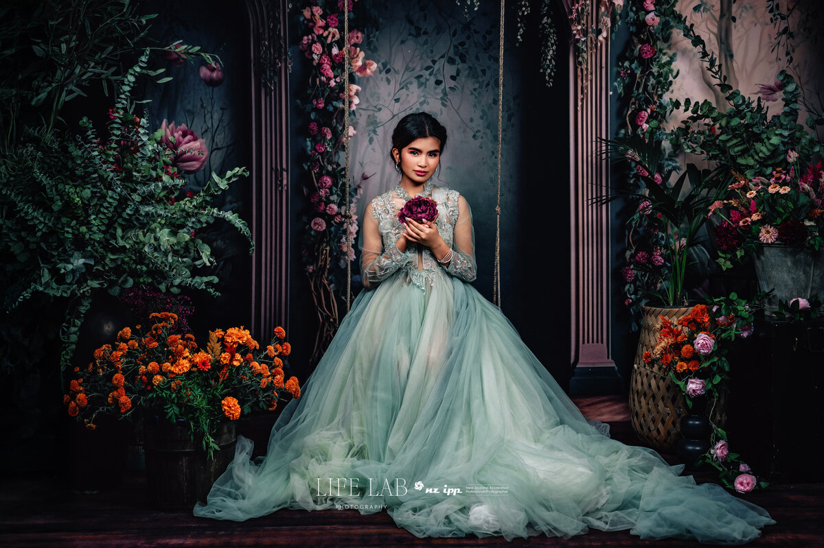 lady sitting in a flower filled room in an extravagant green dress on a swing holding a flower