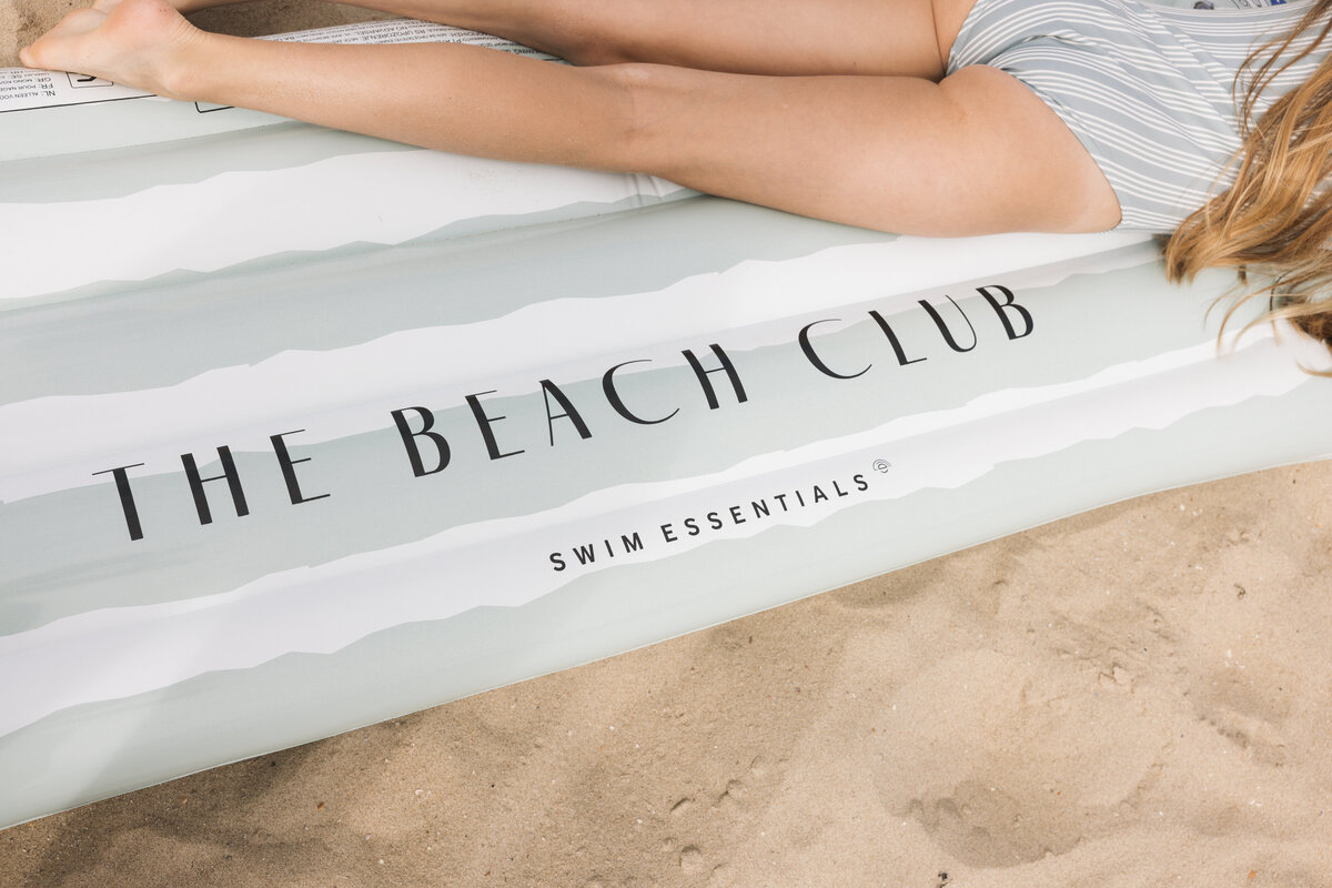 Close-up of a Swim Essentials inflatable air mattress reading “The Beach Club,” with a girl in a striped swimsuit lying on it on sandy beach.