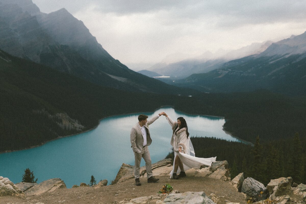 bride and groom dance on rocky outcrop above turquoise lake with mountains in the background