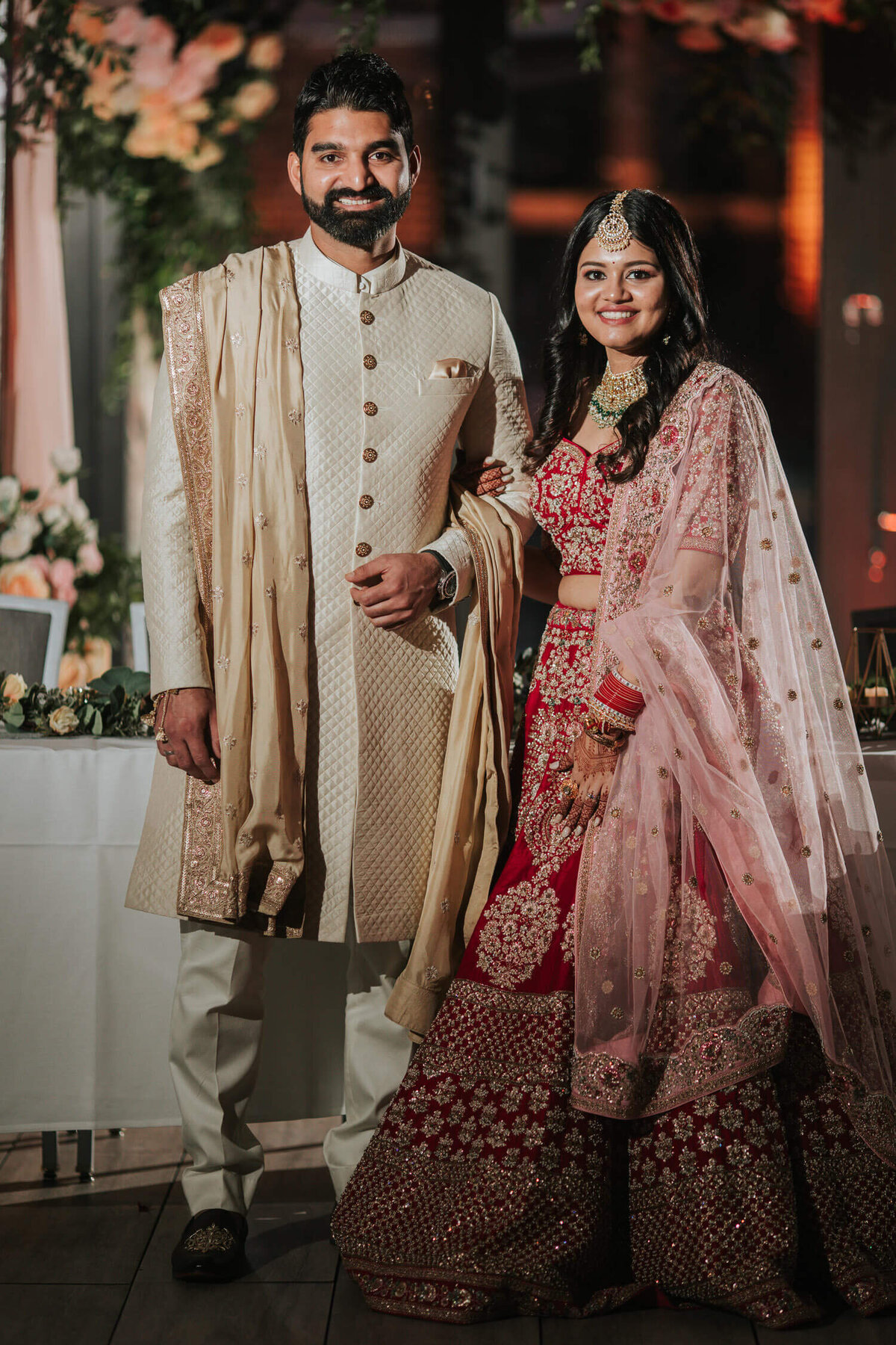 A couple in elegant traditional attire poses at their wedding reception, surrounded by soft floral decor.