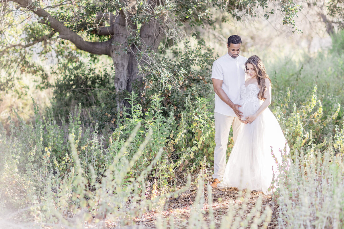 A couple stands in a sunlit forest, surrounded by greenery. The woman is wearing a white dress and holding her pregnant belly, while the man stands beside her, gently embracing her. Sunlight filters through the trees, creating a serene atmosphere.