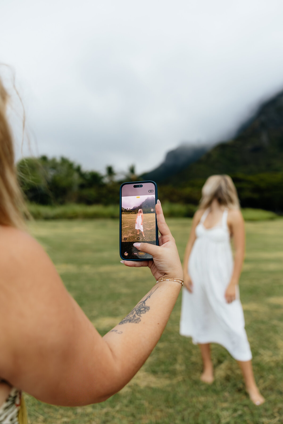 Photographer capturing a friend in a white dress using an iPhone during a photoshoot surrounded by mountains in Oahu, Hawaii.