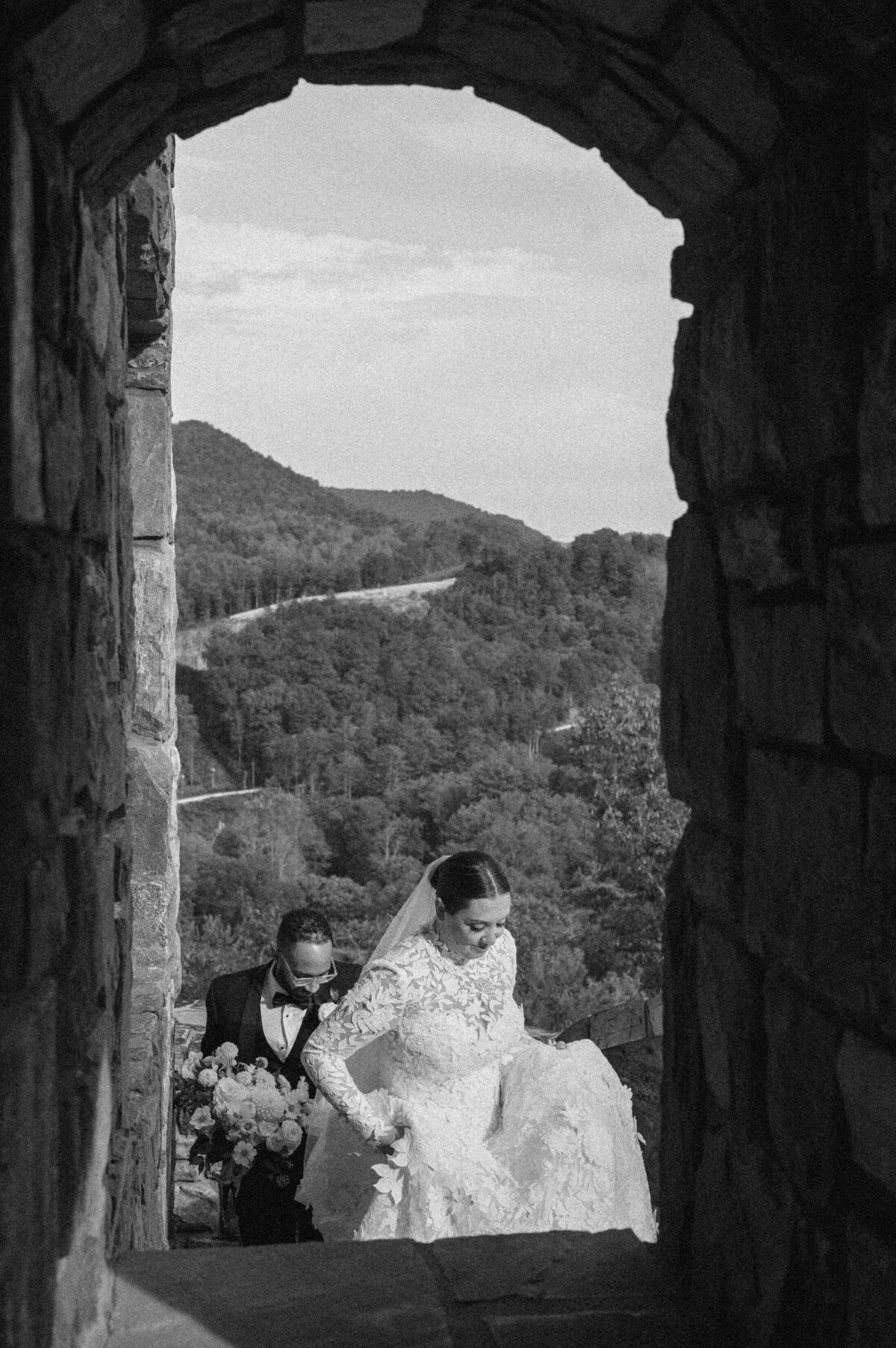 Black and white photo of bride and groom walking through a stone archway with dramatic mountain landscape behind them.