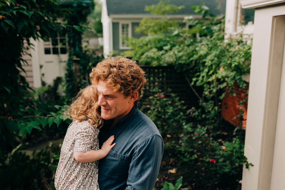 A father with red hair wearing a blue shirt holds his young daughter outside on their back deck