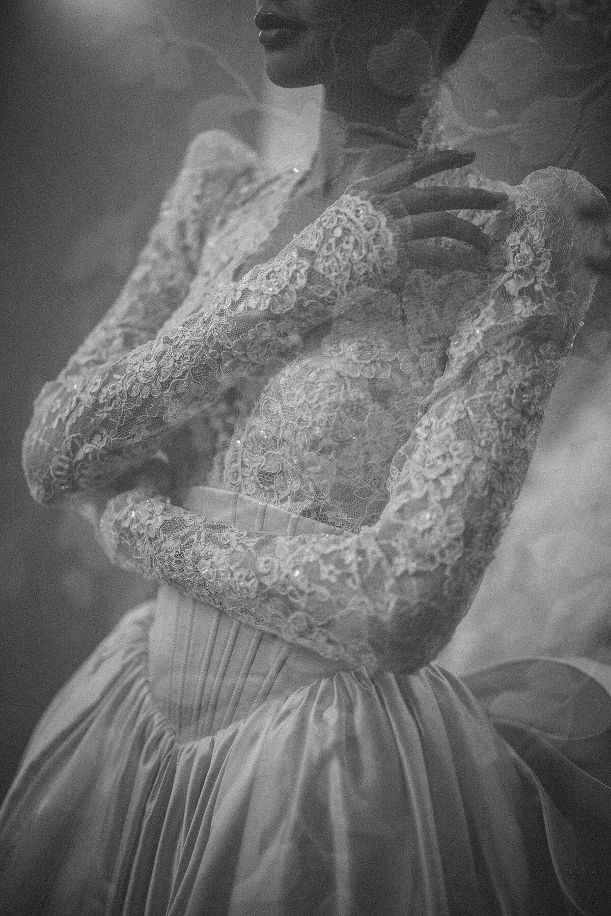 Black and white close-up of a wedding gown featuring intricate lace sleeves, corset detailing, and pleated silk skirt, with soft focus highlighting texture and craftsmanship—an editorial bridal fashion image showcasing timeless couture design.