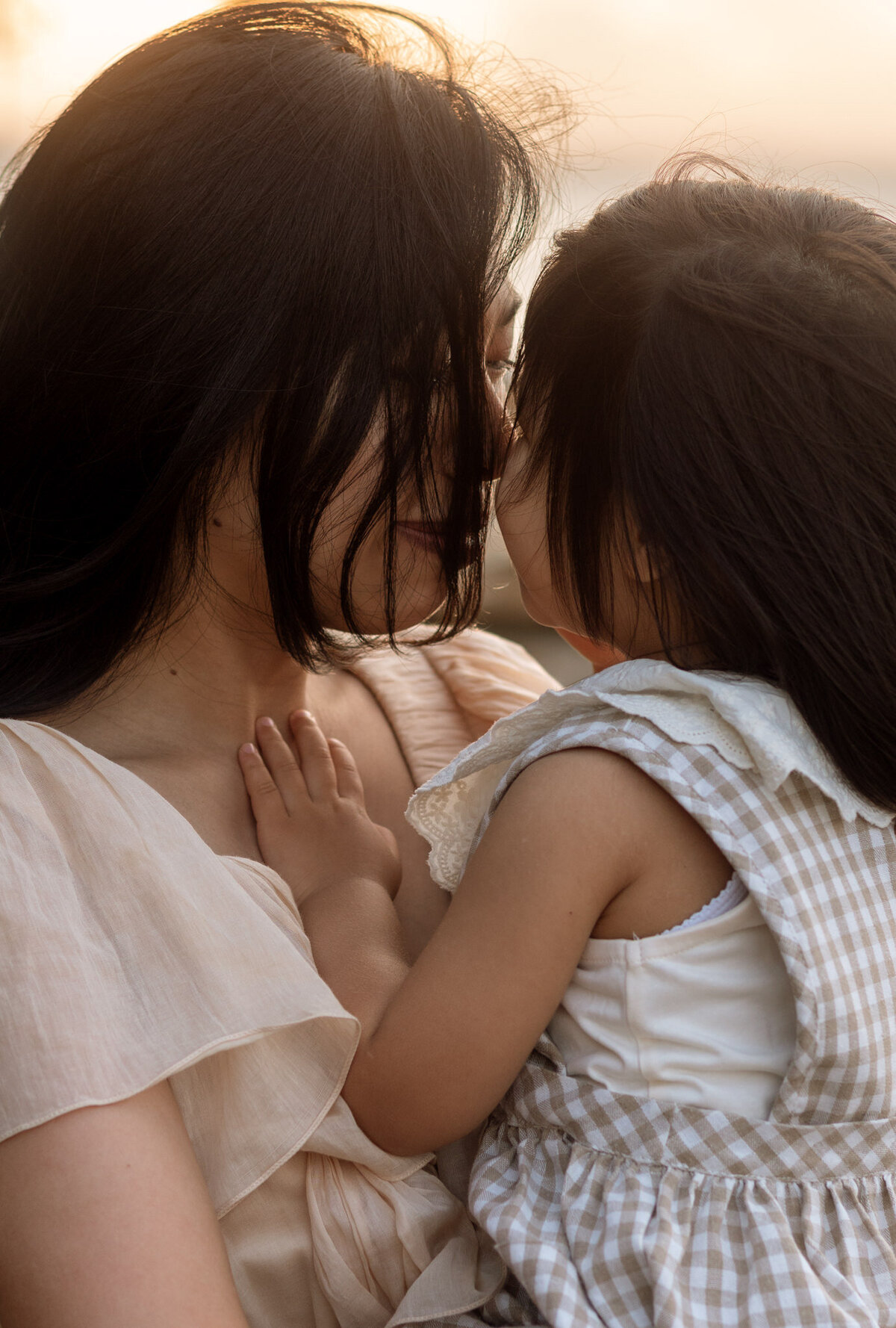 Tender close-up of a mother and daughter sharing a quiet moment, foreheads touching as soft sunlight glows behind them during a family session.
