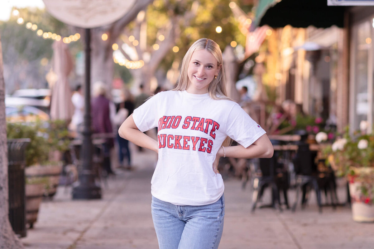 A person wearing a white Ohio State Buckeyes T-shirt poses smiling on a charming street lined with trees and small shops. The street is decorated with lights and has a cozy, inviting atmosphere.