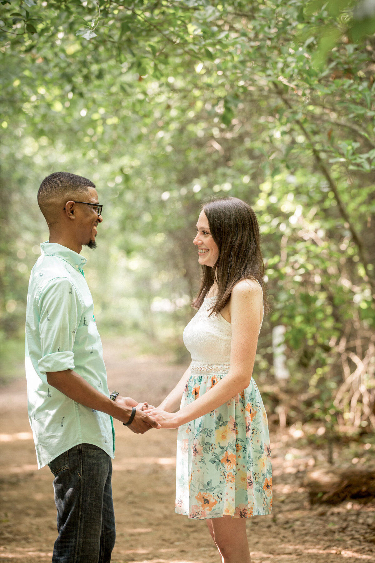 An engaged couple pause to  hold hands and smile at each other during a walk in the springy woods