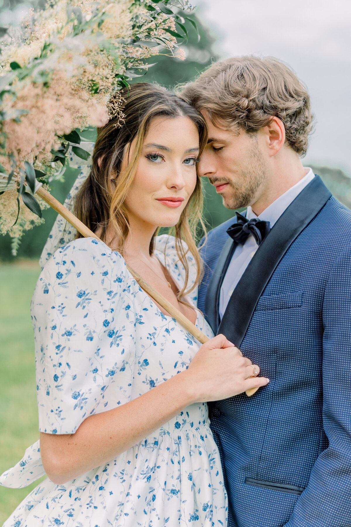 Ivy Rose Barn | A romantic bride and groom embrace in a sunlit field, surrounded by rolling green hills. The bride wears a vintage-inspired floral gown with puff sleeves and a bow and holds a parasol made of flowers, while the groom, in a navy tuxedo, holds her close. Captured by a Richmond wedding film photographer.