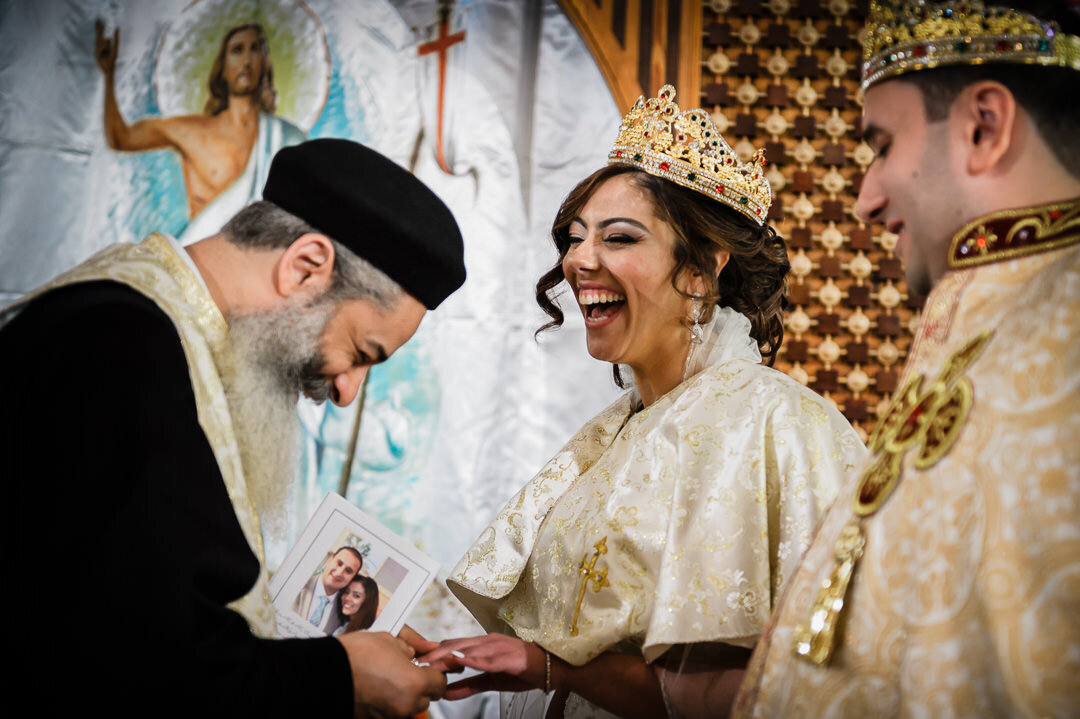 Bride smiles joyfully during Coptic Orthodox wedding ceremony, wearing a gold crown and traditional robe as the priest performs the ring blessing.