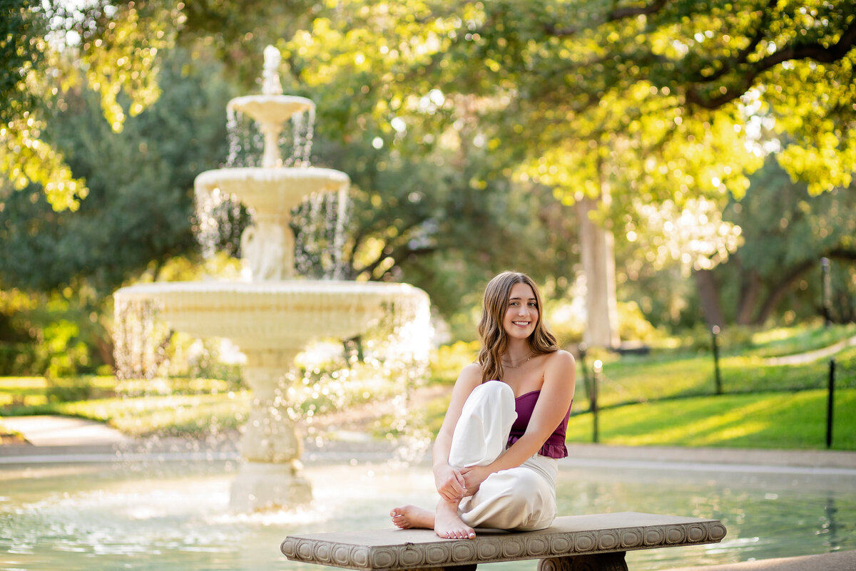 A senior girl poses in front of a beautiful park fountain while sitting on a bench.