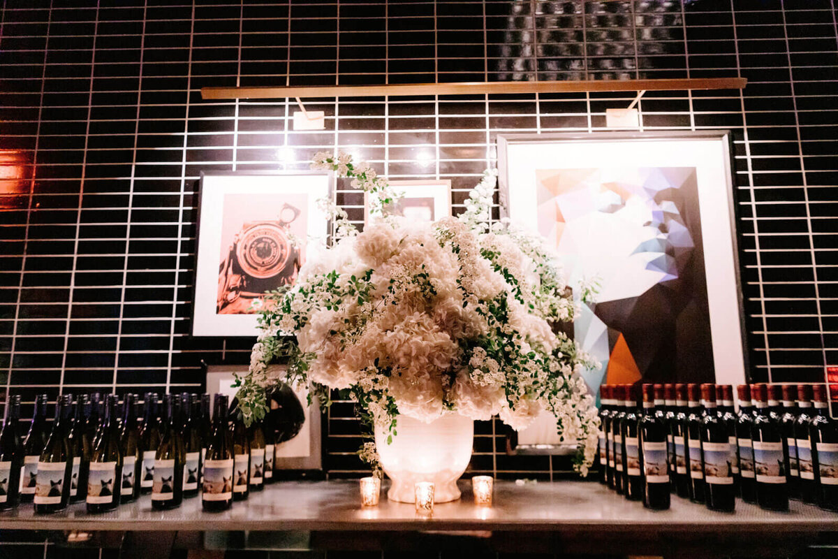 Bottles of champagnes, as wedding giveaways, on a table with a large white flower centerpiece in The Skylark, NYC. Image by Jenny Fu Studio