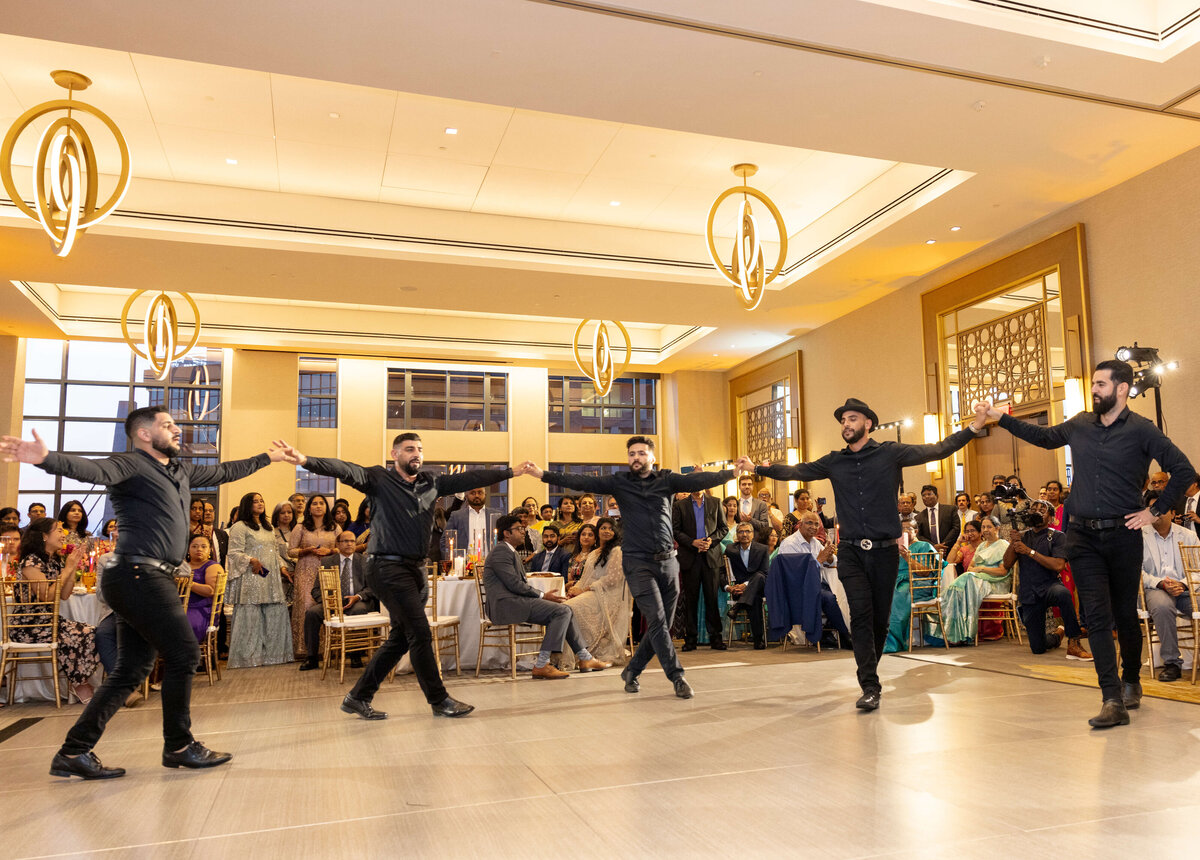 Five men in black outfits perform a traditional dance in a spacious, elegant ballroom. They hold hands in a circle with outstretched arms. An audience sits and watches attentively in the background.