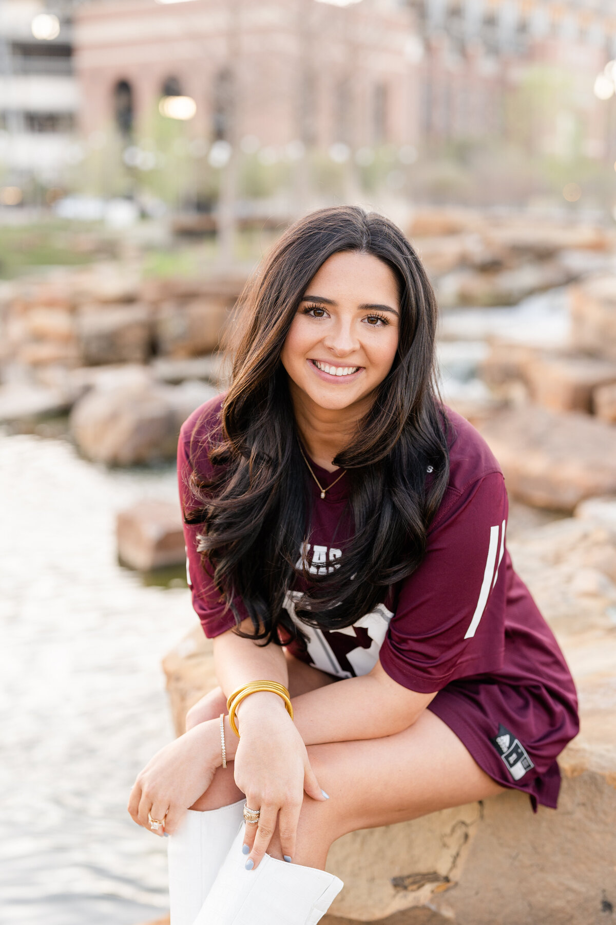 Texas A&M senior girl sitting on rocks and leaning on leg and smiling while wearing Aggie jersey and white boots in Aggie Park
