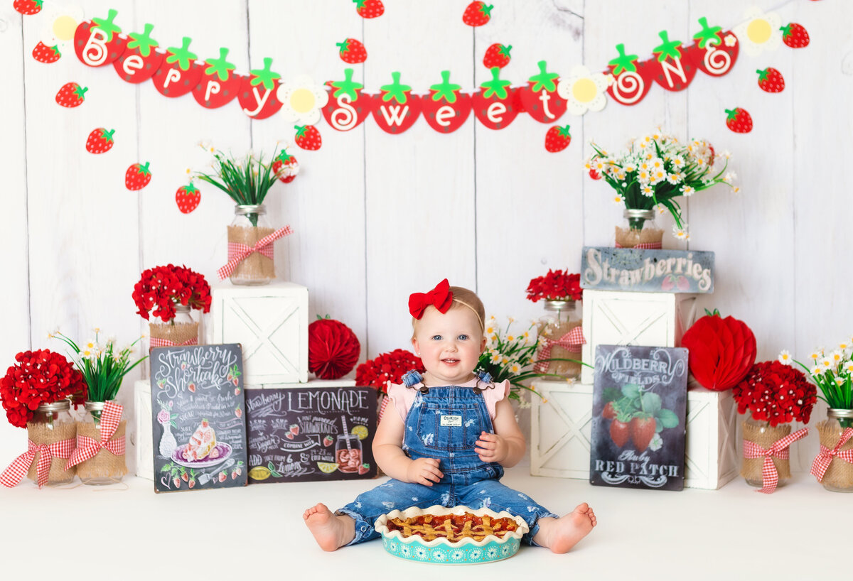 Cake Smash Photographer, a baby sits smiling with overall before a strawberry pie