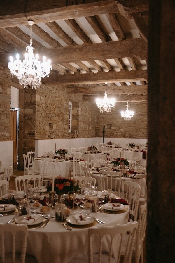 Salle de réception en pierres avec poutres apparentes et tables élégamment dressées sous des lustres étincelants. Mariage capturé par Kamescop en Bretagne.