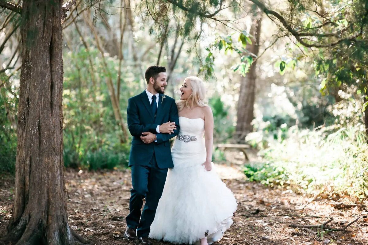 A bride and groom with their arms linked walking in a wooded area.