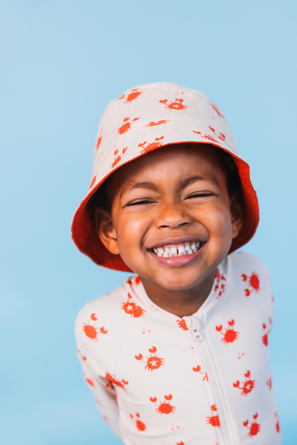 Smiling child wearing a Swim Essentials crab-print UV suit and matching bucket hat against a blue background.