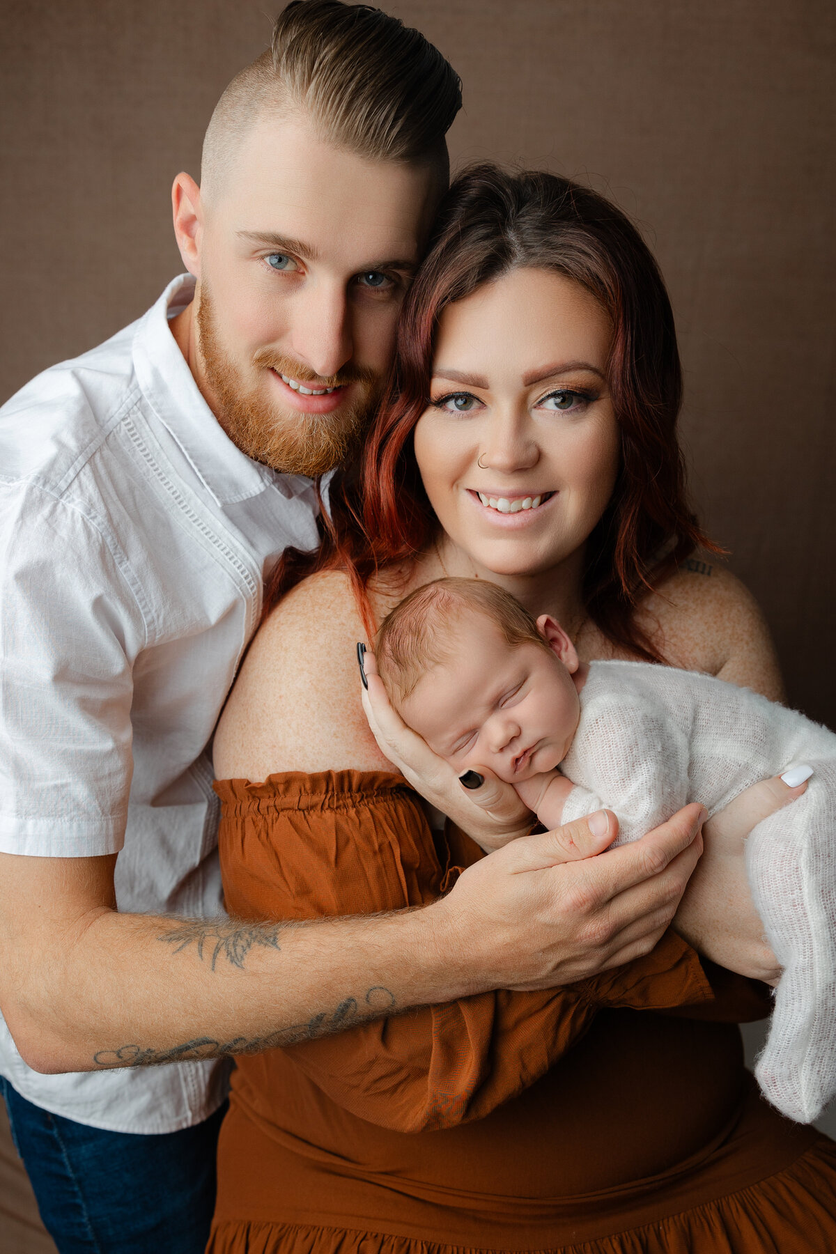 New parents smiling while holding their swaddled baby girl during an in-studio newborn session with a warm brown backdrop. Photograph by Inland Empire newborn photographer Kaitlyn Dawn Photography.