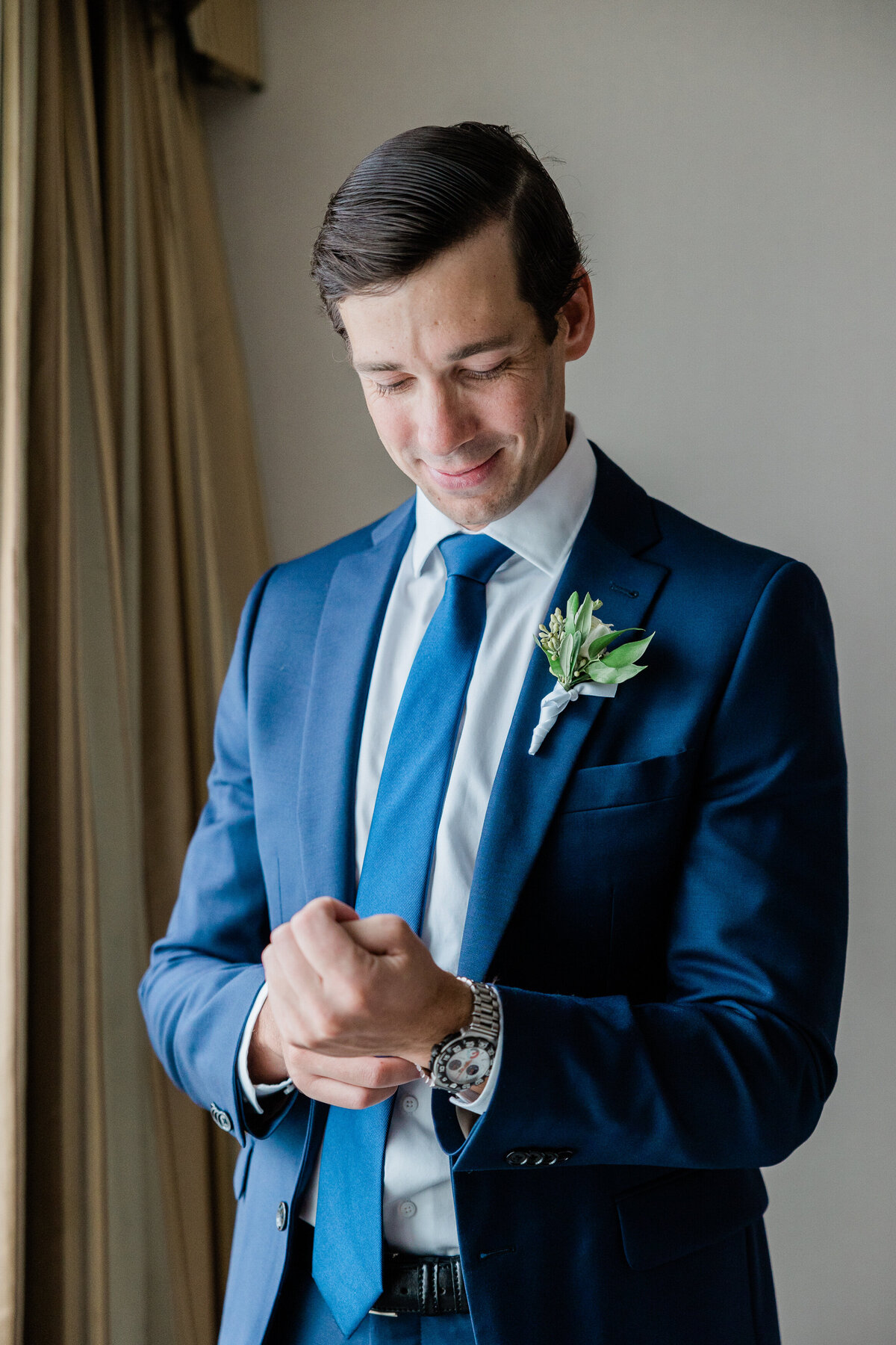 The groom, dressed in a navy blue suit, smiles as he fastens his cufflinks near a window.