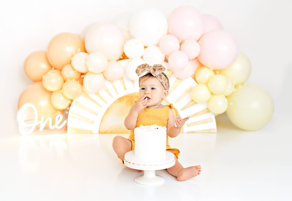 A baby in a yellow outfit sits in front of a white cake on a stand, with a pastel balloon arch in the background. The baby is touching the cake, creating a joyful and playful atmosphere.