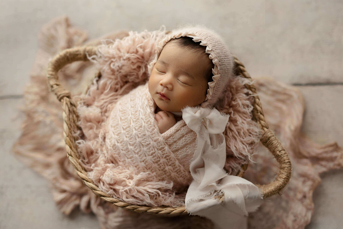 newborn baby girl in a pink swaddle laying in a basket on a white wood floor  with a pink bonnet with bow ties at a newborn photo shoot in Northern VA