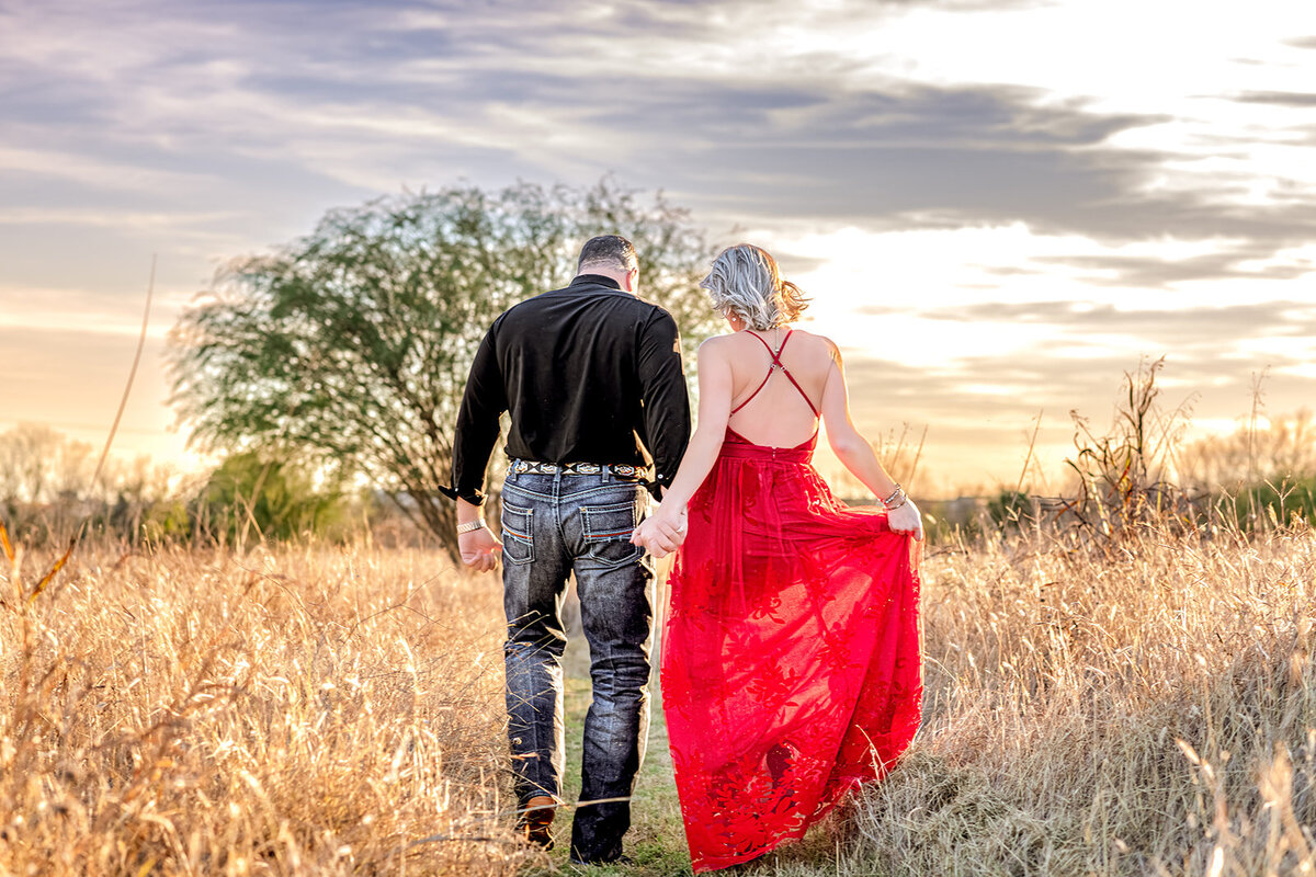 Engaged couple walking away from camera in field at sunset during engagement session in New Braunfels, Texas. Bride in red dress, groom in jeans and cowboy hat.