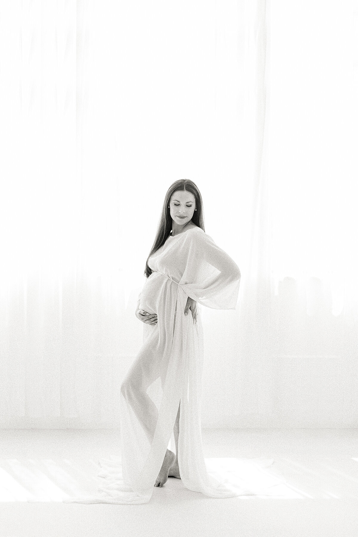 Black and white portrait of a pregnant mother posing in a DFW photography studio for her maternity session as she is wearing a chiffon gown as she holder her belly and looking over her shoulder.