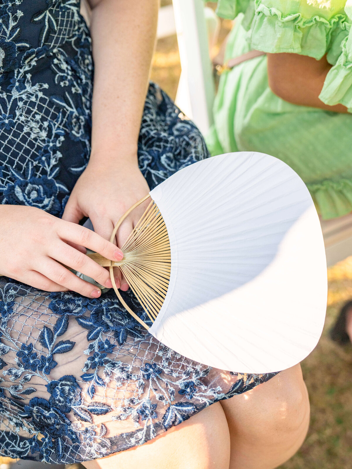 Two people seated outdoors, one holding a white paper fan with a bamboo frame. The person on the left wears a blue lace dress, while the one on the right is in a light green ruffled outfit. Sunlight highlights the textures and colors.