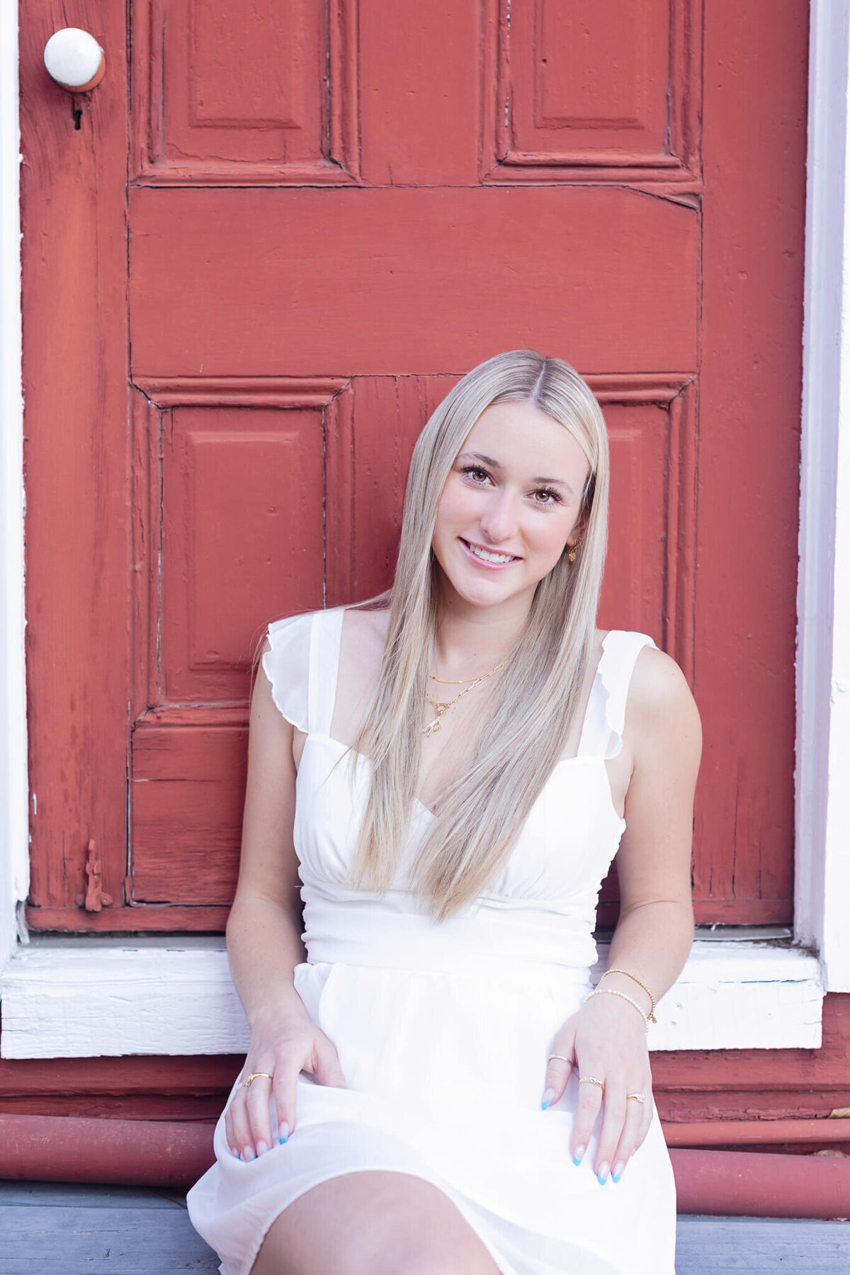 A person with long blonde hair sits in front of a red wooden door, wearing a white dress. They are smiling and have their hands resting on their lap.