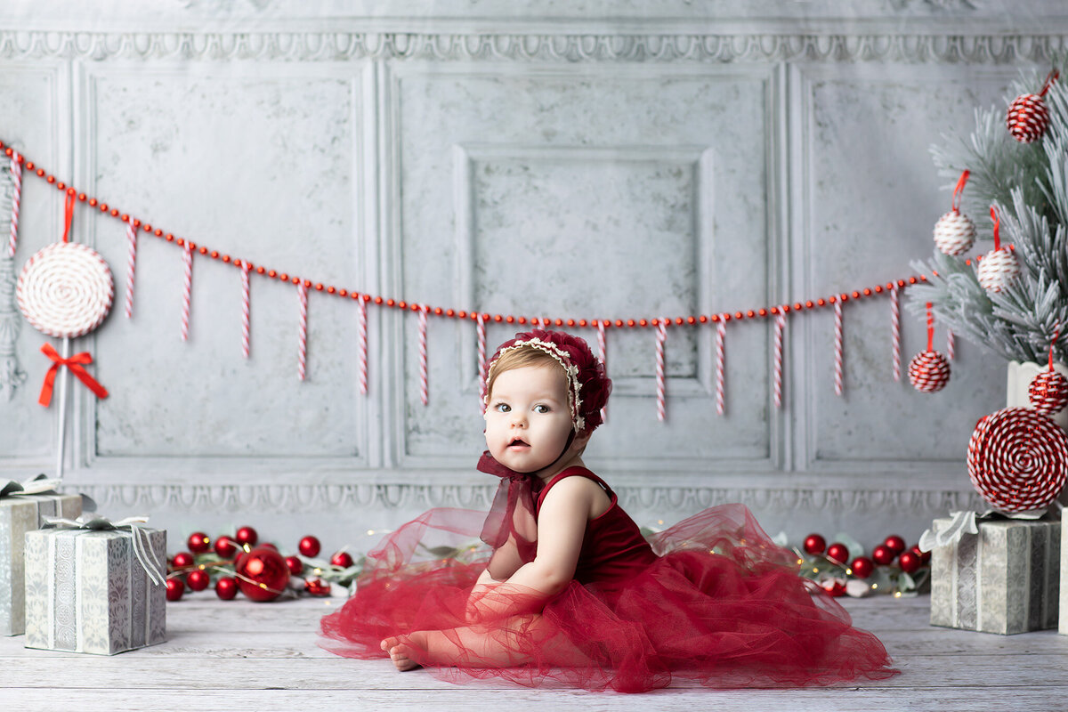 Baby girl in a red tulle dress sitting on a wood floor surrounded by candy cane garlands, presents, and peppermint decorations, photographed by a DFW milestone photographer.