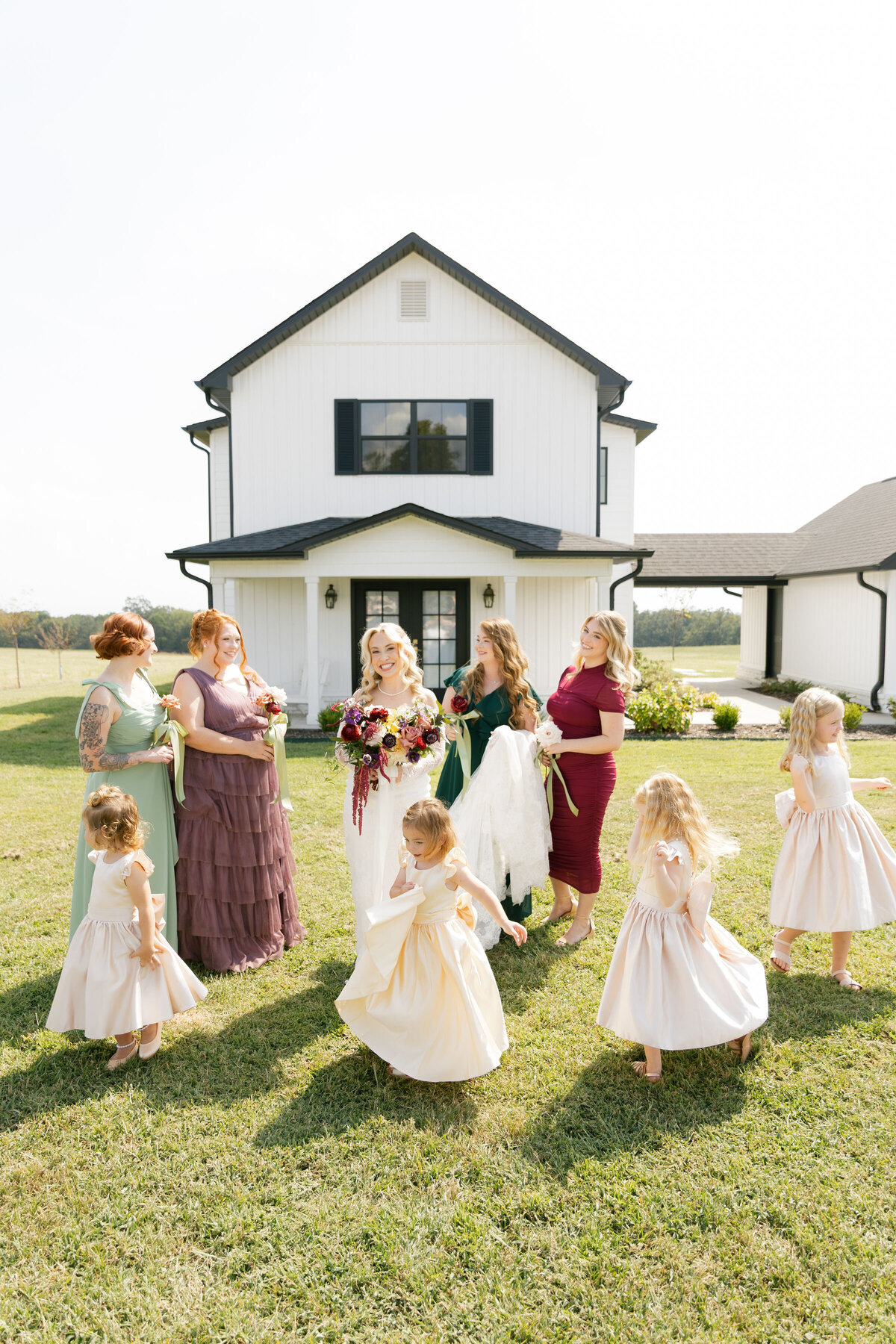 Bride and bridesmaids laughing together in front of a modern white farmhouse wedding venue, accompanied by young flower girls twirling in cream dresses. Bridesmaids wear mismatched jewel-tone gowns and hold single-stem roses with sage ribbons, creating a whimsical, joyful wedding morning moment.
