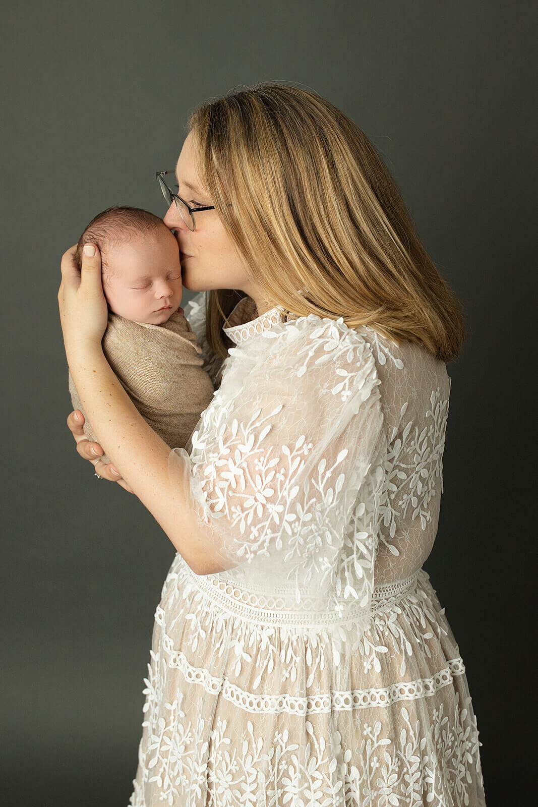 mom kissing the side of her newborn baby boy's head on a dark grey background
