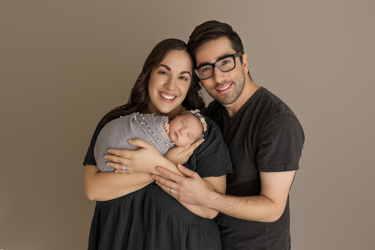 Smiling parents holding their sleeping newborn baby wrapped in a grey blanket, standing against a neutral beige background.