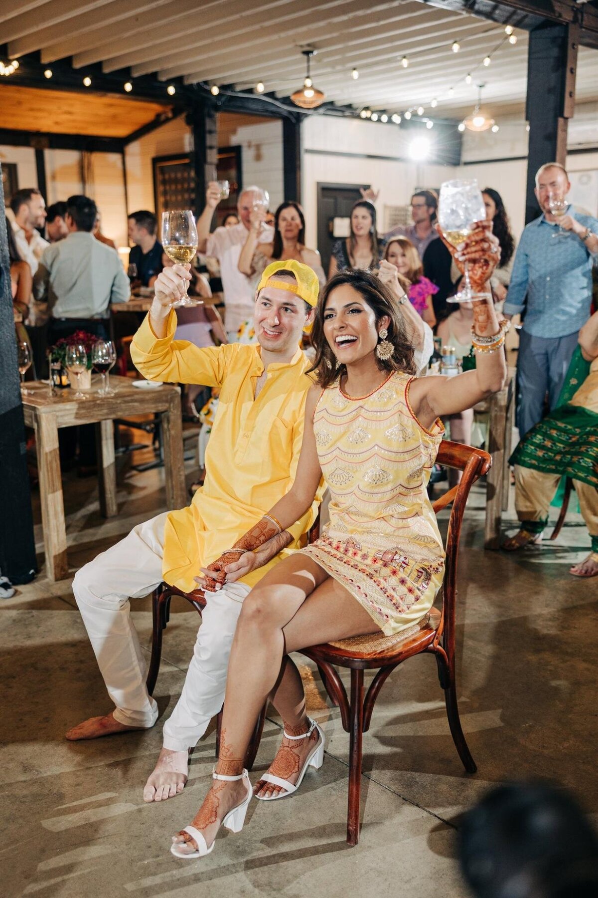 A couple in traditional attire sitting and raising glasses at a lively event with guests in the background.