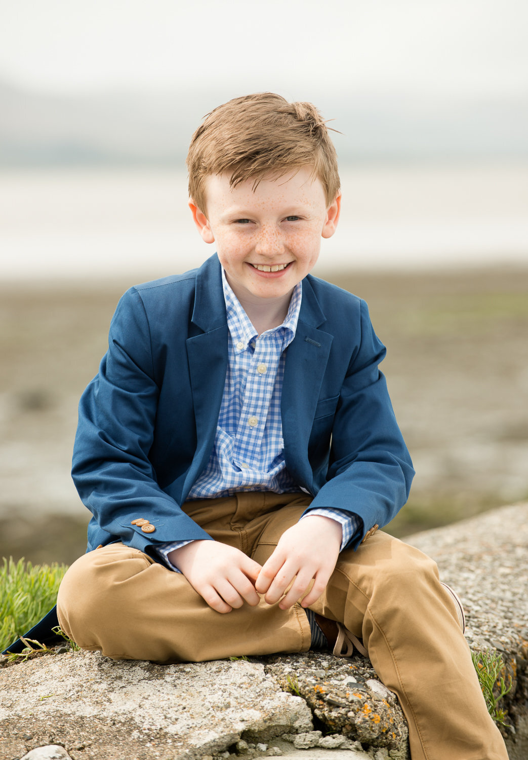 boy in a blue blazer and brown chinos sitting on a wall, smiling at camera