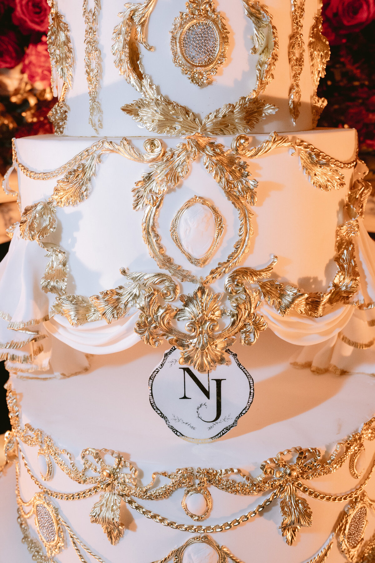 The photo was taken at Shangri La Hotel & Ritz Paris Hotel in Paris, France by photographer Matteo Coltro and videographers Zen Film Works. It captures a close-up of an elaborate, multi-tiered white wedding cake. The cake is adorned with intricate gold embellishments and decorative patterns, including ornate garlands and leaf accents. Prominently displayed on one tier are the initials N and J within a crest, adding a personalized touch to the stunning confection.