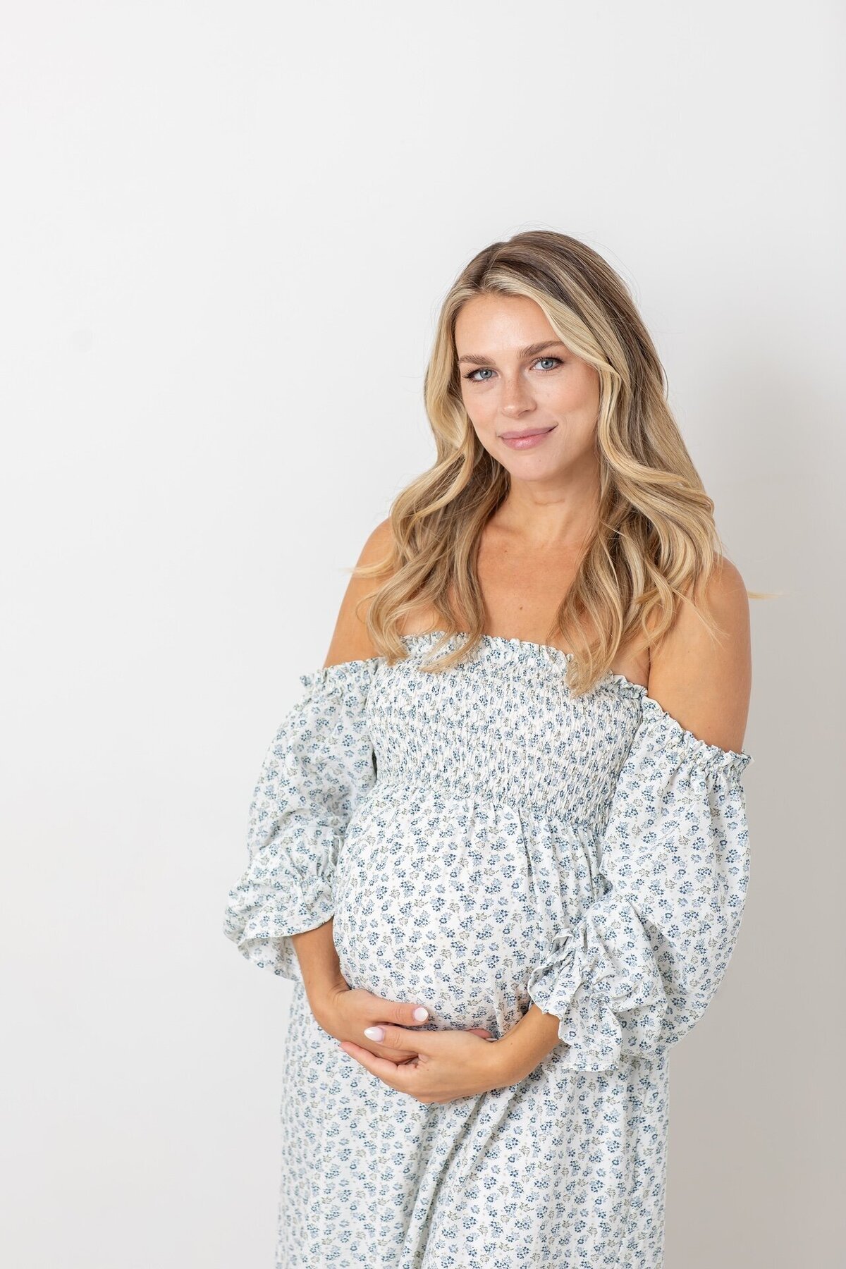 Maternity portrait of a woman wearing a white and blue dress, holding her baby bump with a peaceful expression