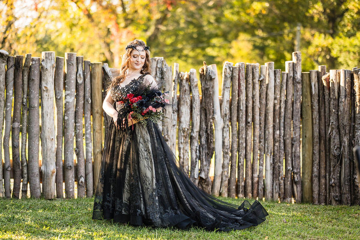 A lesbian bride wearing a black wedding dress stands in the backyard of an Airbnb at golden hour in Roanoke, Virginia for a formal portrait. The bride looks over her shoulder at a bouquet that she made herself.