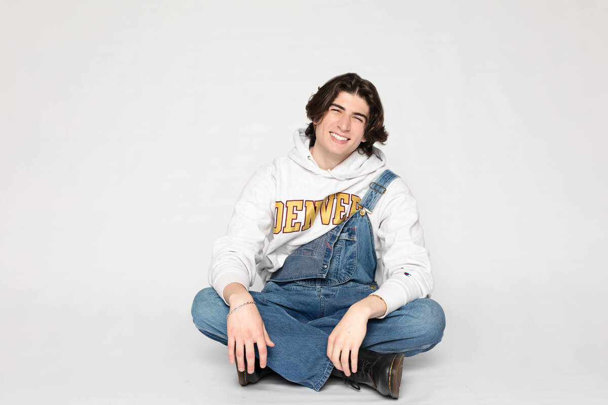 A person with shoulder-length hair sits cross-legged on the floor, wearing a white hoodie with Denver on it and blue denim overalls. The background is plain white, and they are smiling at the camera.