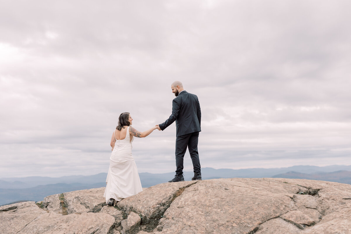 At the peak of a mountain a man helps a woman climb up a rock to see the view of Lake George during their elopement.